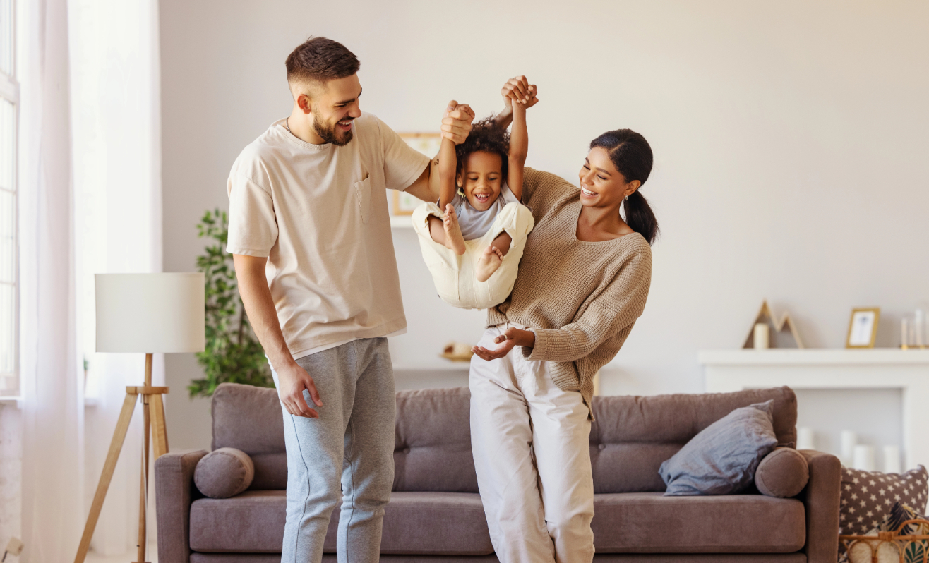 Full body multiethnic father and mother smiling and lifting mixed race son near couch in cozy living room